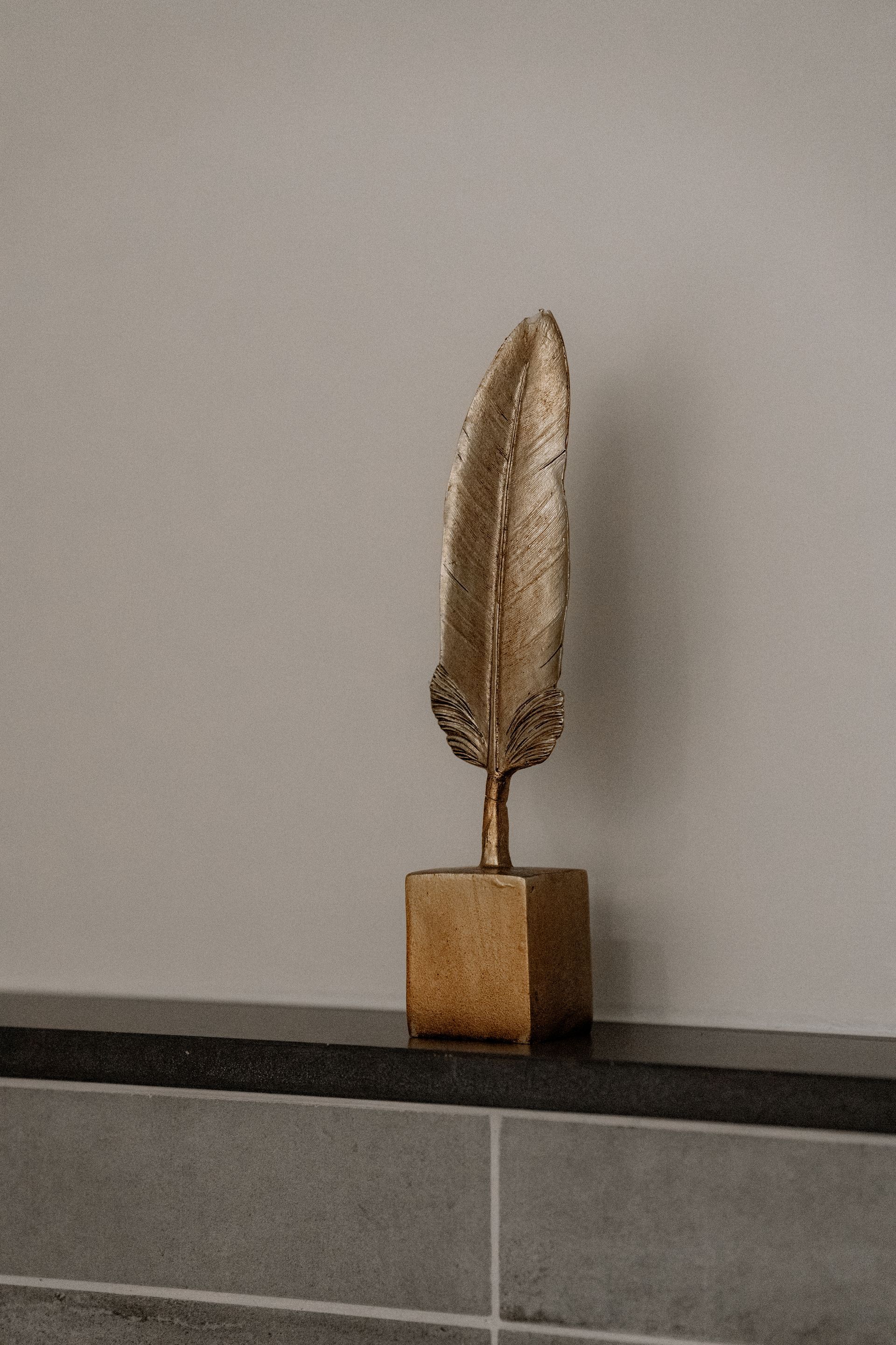 Een sculptuur van een gouden veer op een vierkante sokkel, die op een donkere plank tegen een grijze muur staat.