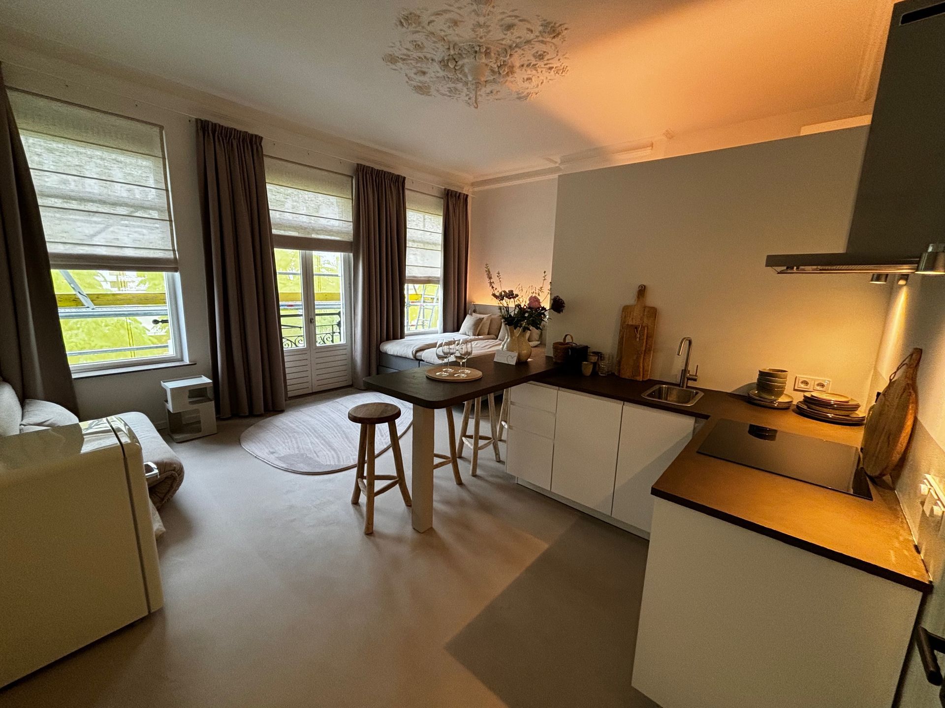Gezellig studioappartement met een bed, een kleine keuken en een zithoek bij grote ramen met neutrale gordijnen.