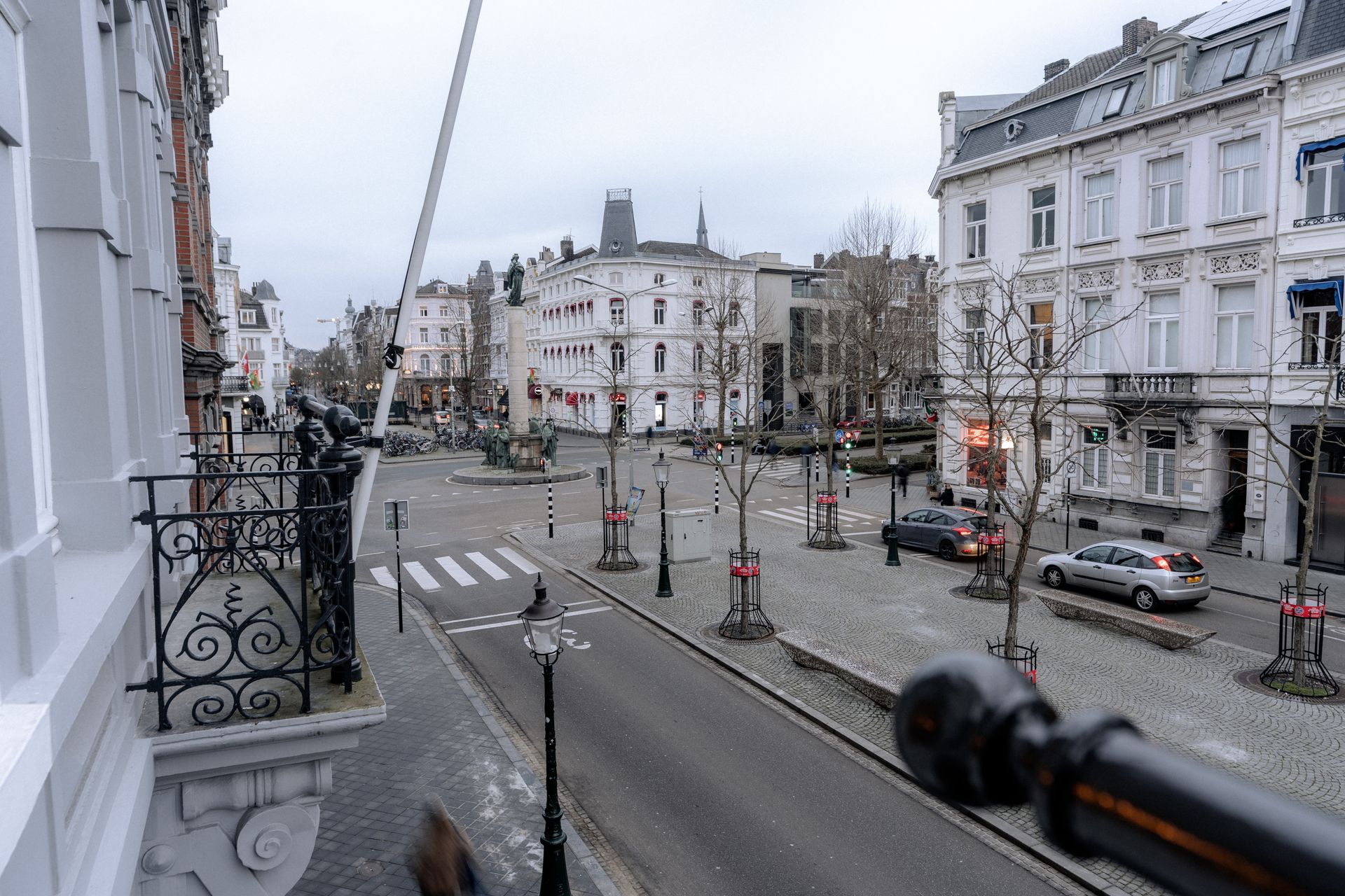 Straatbeeld: geplaveide weg, gebouwen, auto's, kale bomen en een bewolkte lucht.