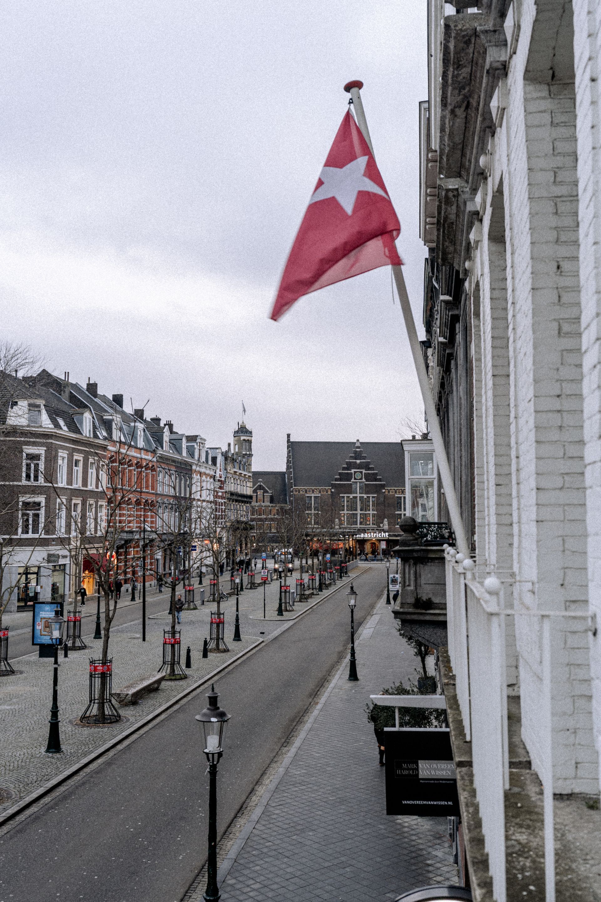 Een straatbeeld met een rood-witte vlag, Europese gebouwen en een bewolkte hemel.