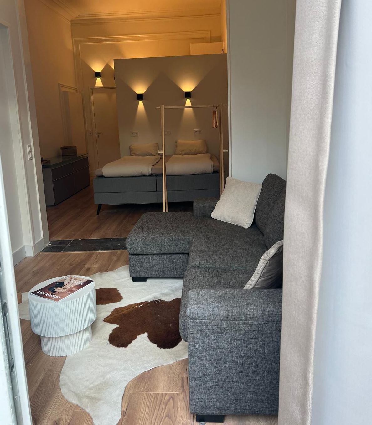 Woonkamer met grijze hoekbank, koeienhuid vloerkleed en uitzicht op een slaapkamer met twee bedden.