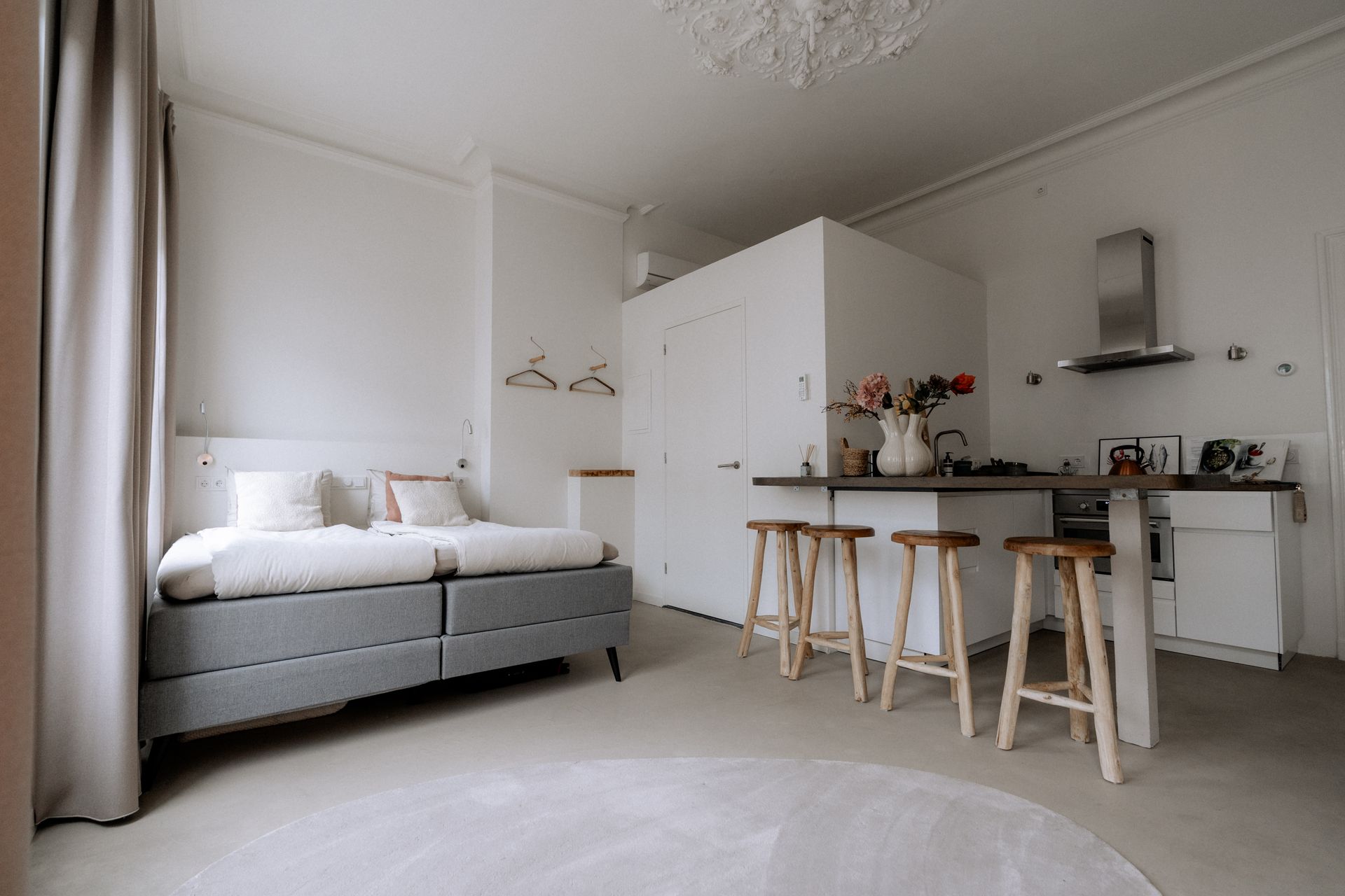 Licht studioappartement met bed, keuken en kookeiland met barkrukken. Witte muren, grijs bed, lichte houten barkrukken.