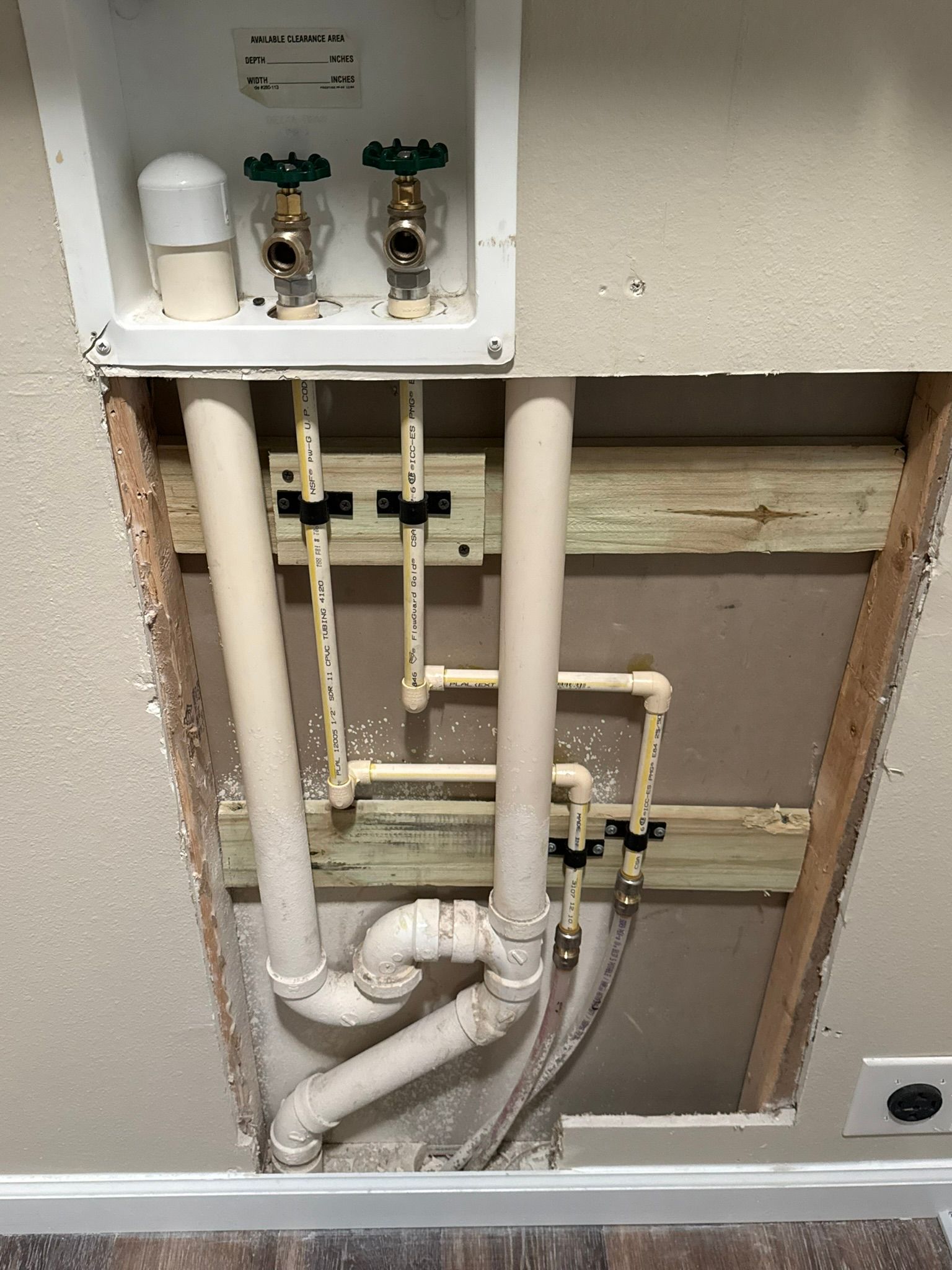 Open plumbing access panel with pipes, valves, and white PVC lines inside a wall niche.