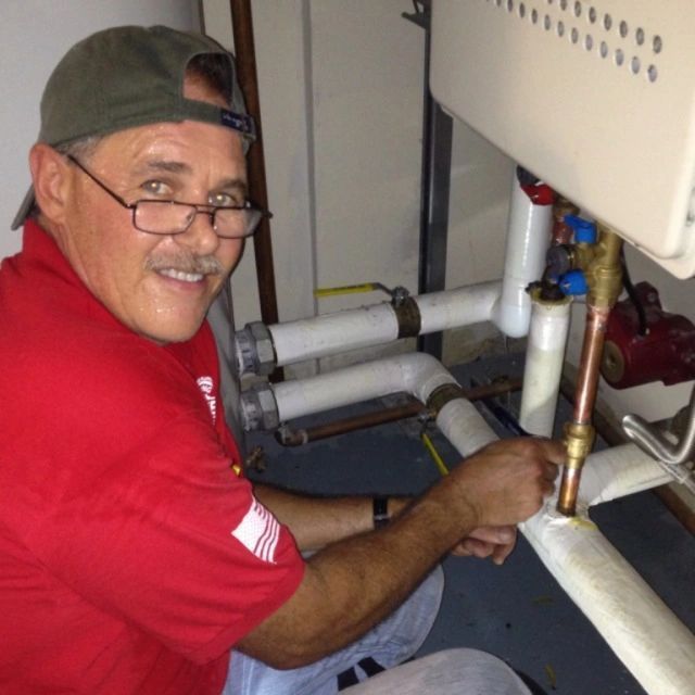Worker in red shirt inspecting plumbing pipes under a sink, smiling at the camera.