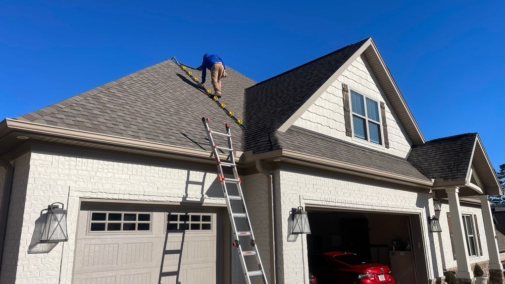 Worker on a ladder repairing a gray shingled roof above a house garage on a sunny day