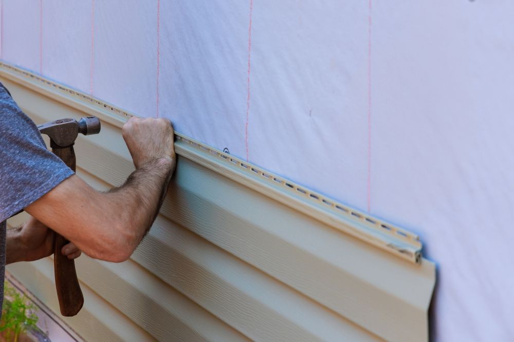 Hands installing beige siding panels on an exterior wall with a hammer and pry bar