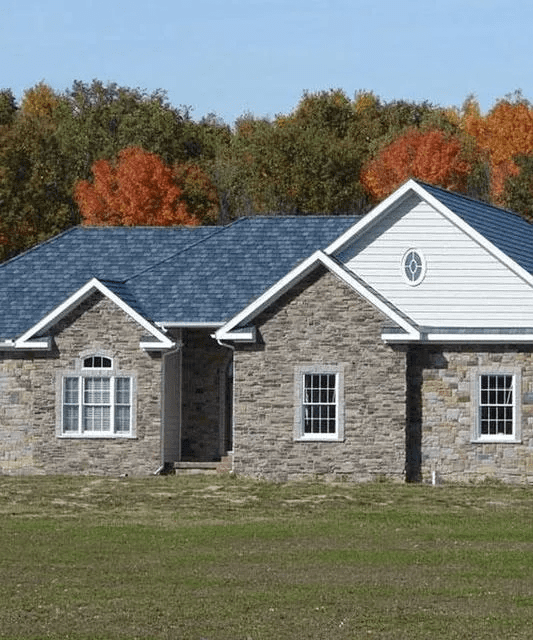 Stone house with gray roof and white siding, set in a grassy field with autumn trees behind.