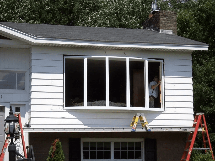 Workers installing large white windows on a house exterior, with ladders on both sides.