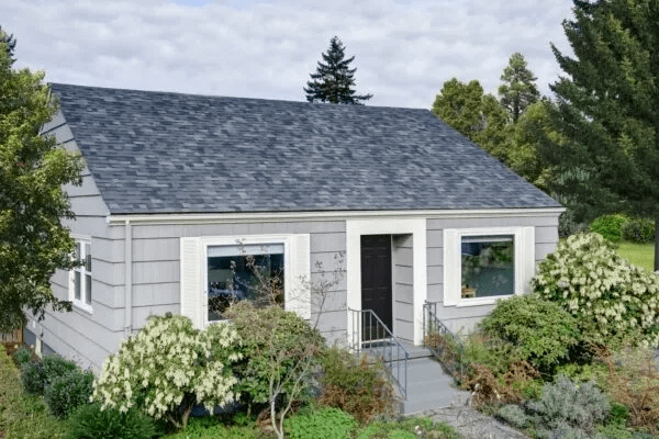Small gray house with a dark roof, front door, and overgrown shrubs in the yard