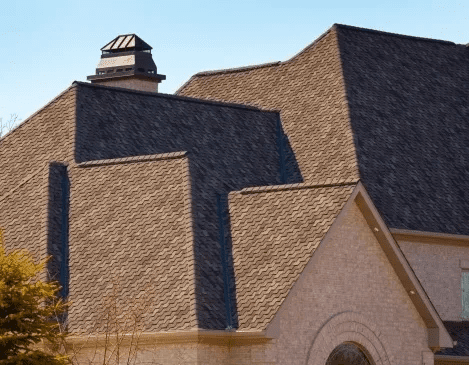 Tan shingled building roof with a small cupola and a stone gable under clear sky