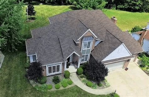 Aerial view of a gray-roofed suburban house with a curved walkway and driveway, surrounded by green lawn and trees