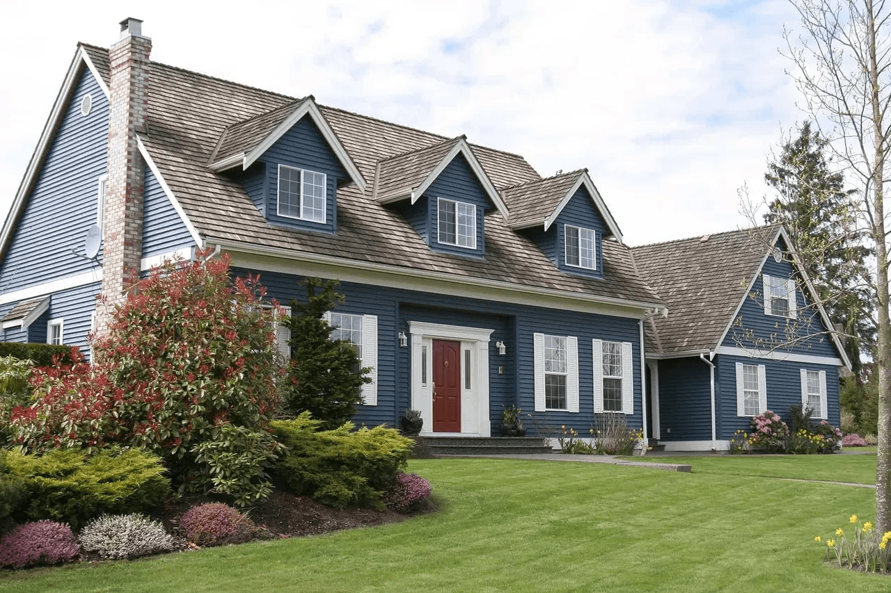 Blue house with white trim, red front door, dormer windows, and a green lawn under a cloudy sky