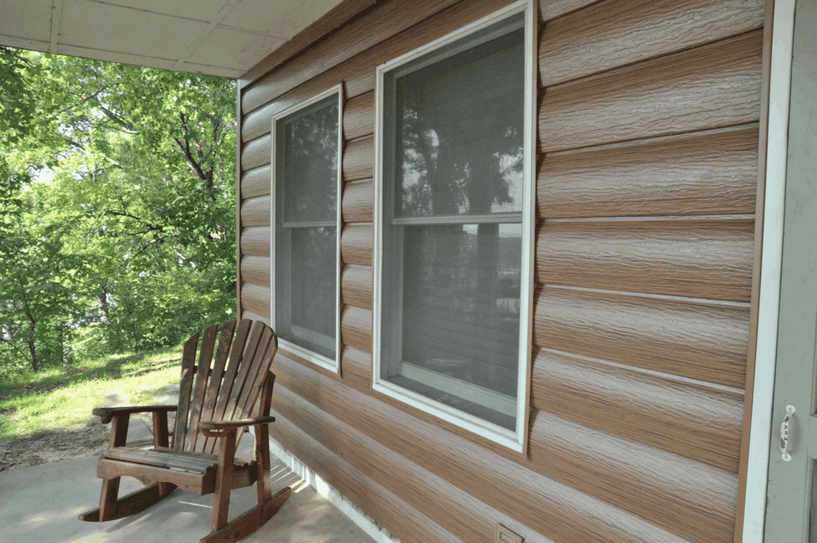 Wooden rocking chair on a covered porch beside tan siding and windows, with a green yard beyond.