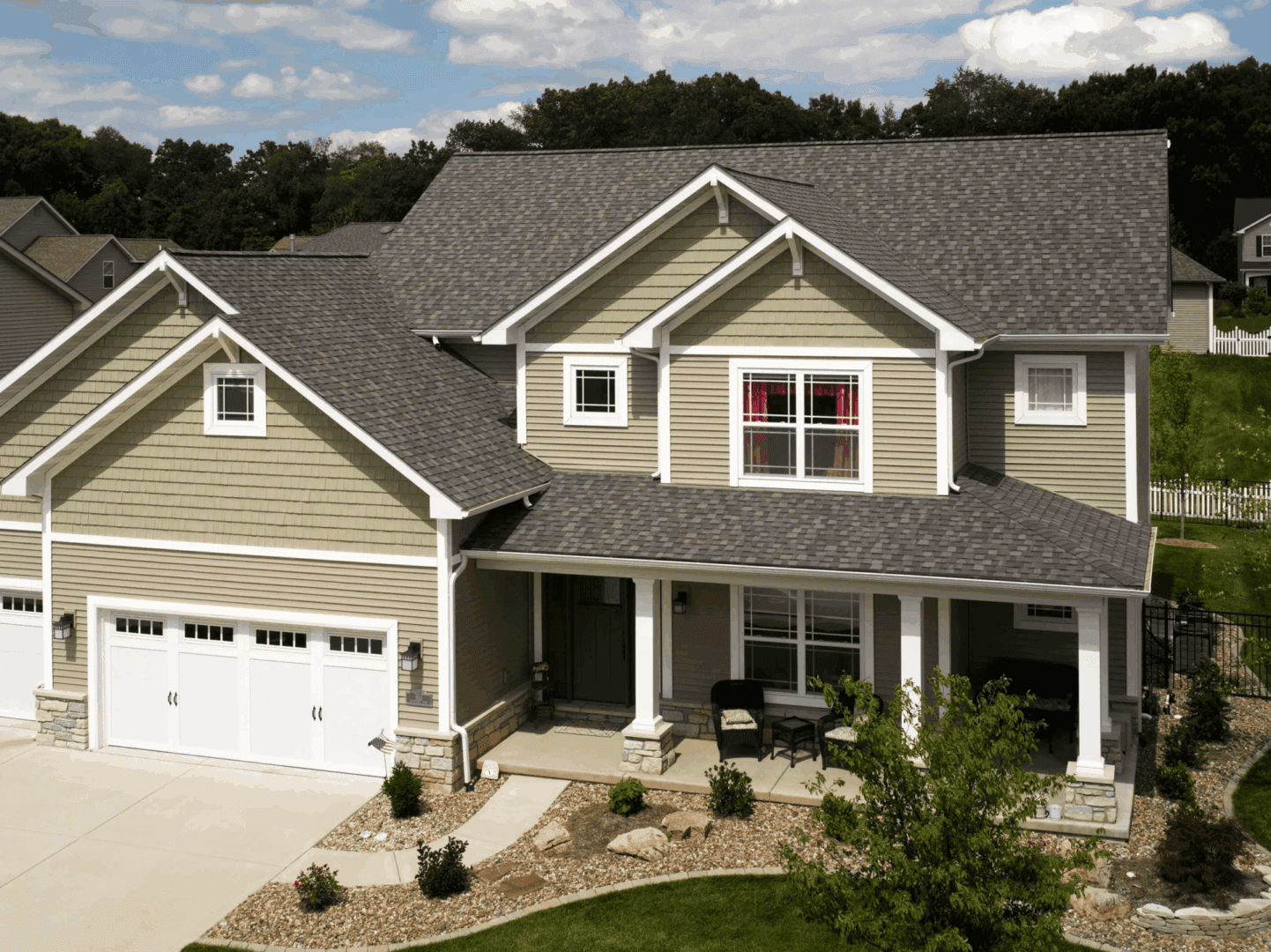 Two-story suburban house with beige siding, gray roof, white garage, and front porch under a blue sky