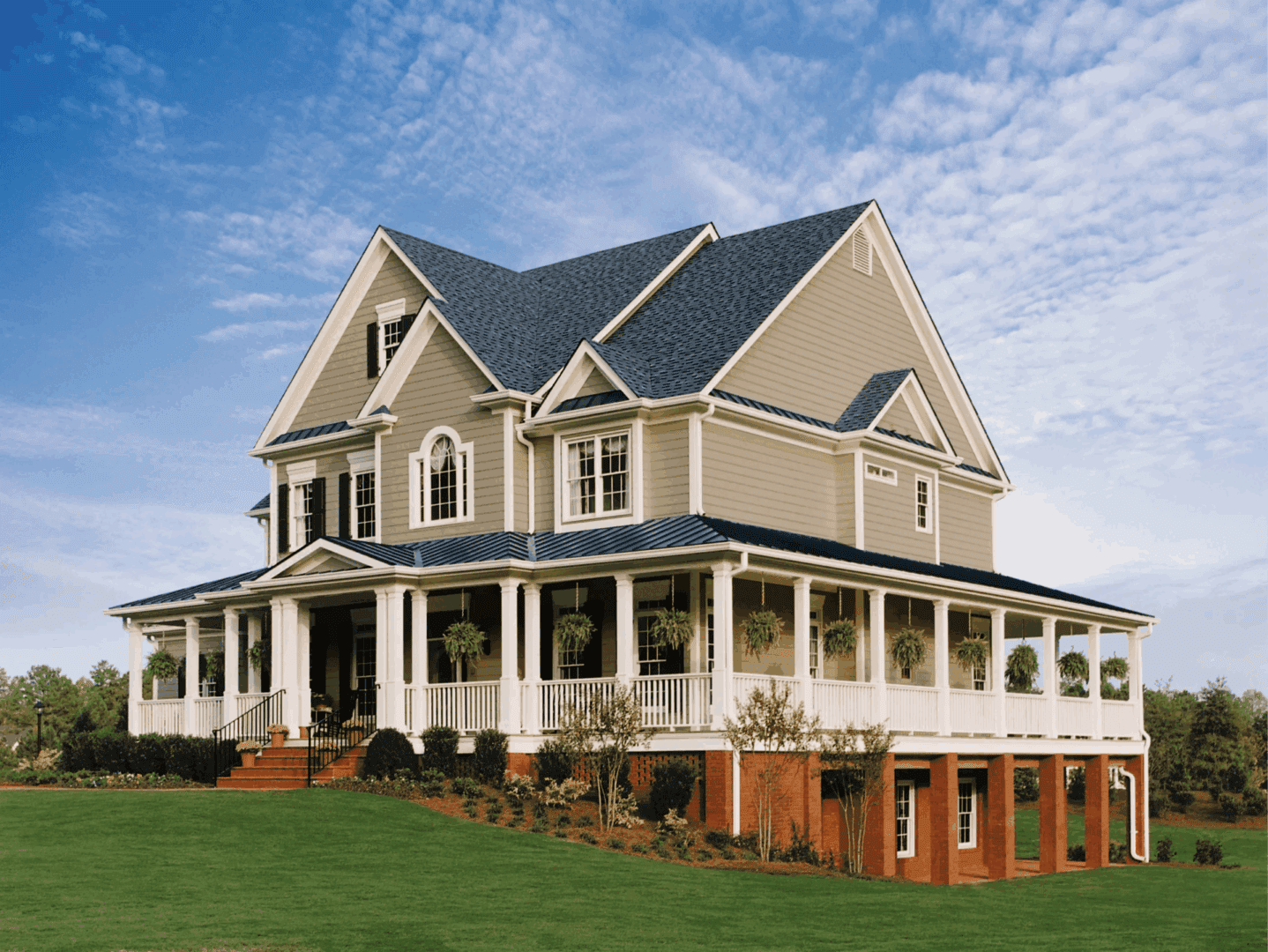 Large beige Victorian-style house with white wraparound porch and dark roof on a grassy lawn under a blue sky