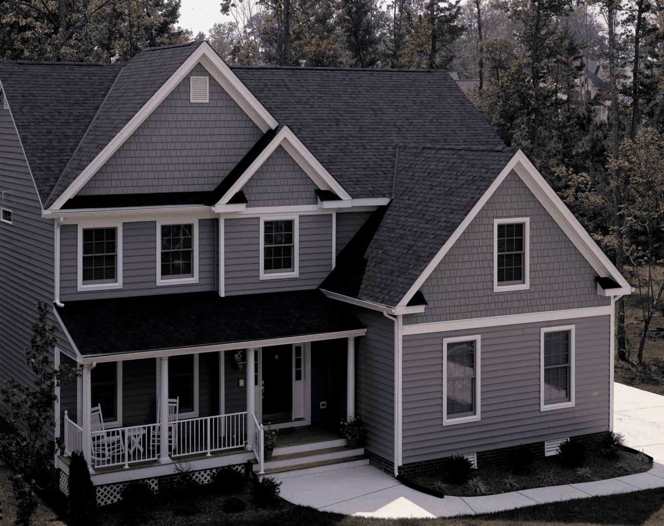 Two-story gray suburban house with dark roof and white porch railings surrounded by trees