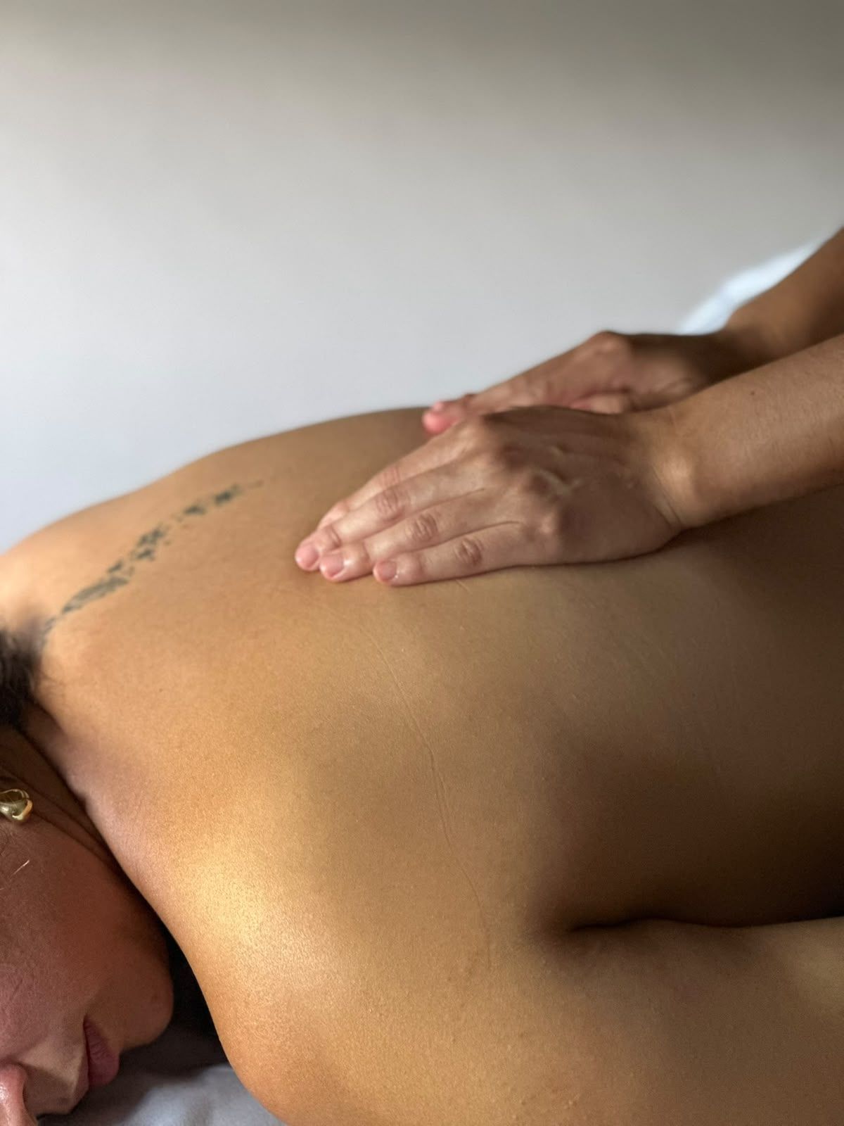 Persona recibiendo un masaje en la espalda, con las manos presionando la parte superior de la espalda desnuda sobre un fondo liso.