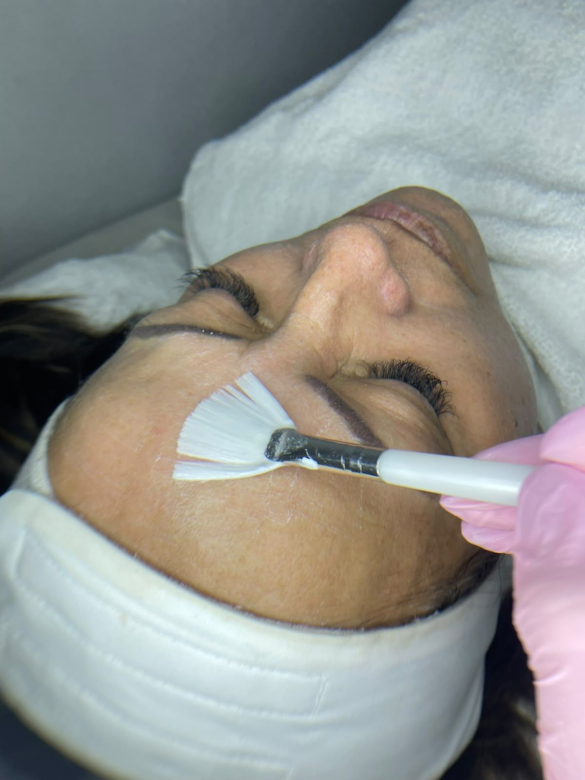Una esteticista aplica una mascarilla facial blanca con un pincel a una clienta recostada con los ojos cerrados