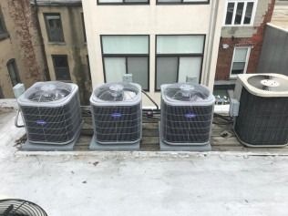 Four rooftop air conditioning units lined up on a flat rooftop beside nearby buildings.