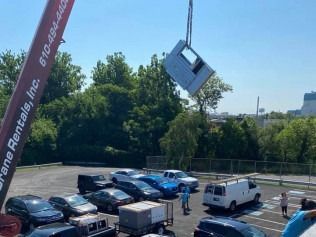 A large suspended blue car hangs over a parking lot with cars, trees, and workers below.