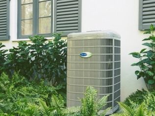 Outdoor air conditioner unit beside a house, surrounded by green shrubs and shutters.