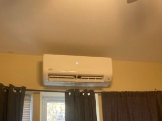 White wall-mounted air conditioner above a window with dark curtains in a beige room