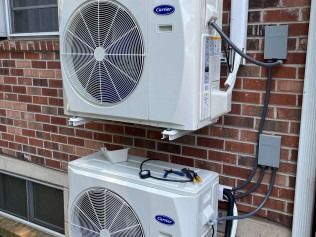 Two white air-conditioning units mounted on a brick wall with electrical boxes nearby