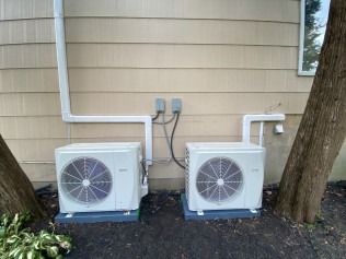 Two outdoor air conditioning units beside a house wall, with trees and utility boxes nearby