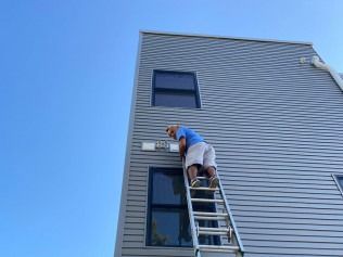 Worker on a ladder painting the exterior wall of a gray house under a blue sky
