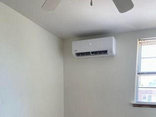 White room with a wall-mounted air conditioner, ceiling fan, and window with blinds.