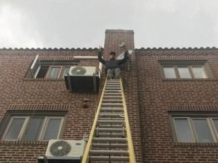 Person standing at top of a tall ladder against a brick building, arms raised overhead