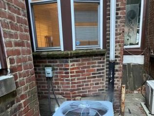 Brick backyard wall with windows, metal AC unit, and outdoor electrical box in a narrow alleyway