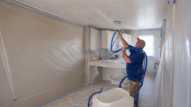 Two workers renovating an interior, one on a ladder repairing a ceiling and one framing a wall.