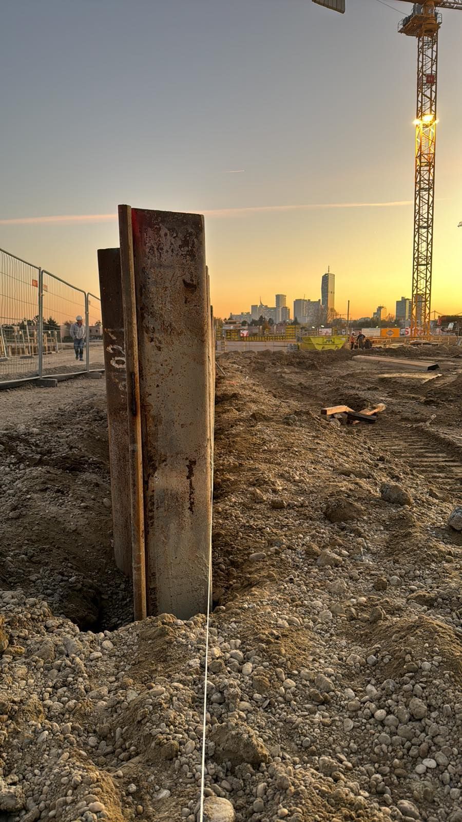 Baustelle mit Stahlpfählen im Vordergrund, Skyline der Stadt und Kran bei Sonnenuntergang.