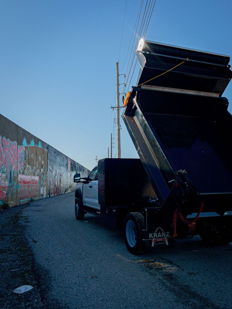 Truck with raised dump bed on a gravel road beside a graffiti-covered wall.