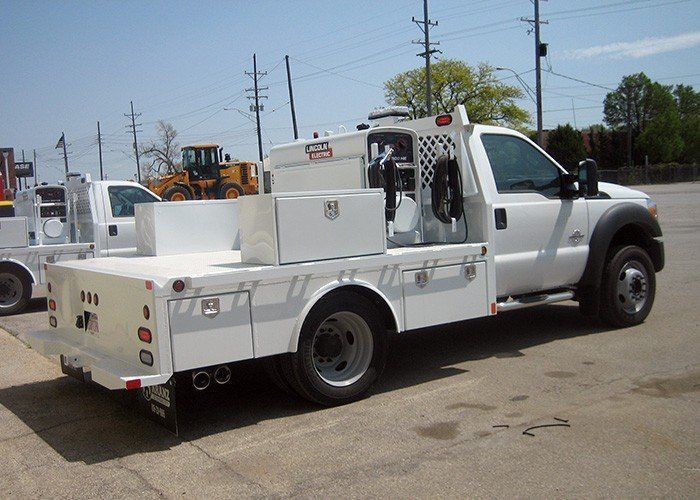 White service truck parked outdoors on a sunny day. A flatbed with storage compartments and equipment is mounted on the truck.