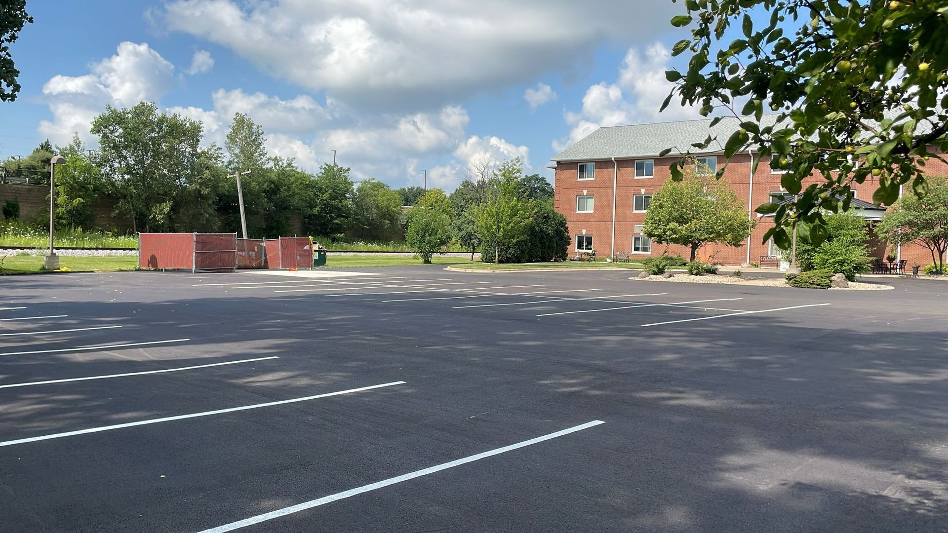 Empty asphalt parking lot with white painted lines; red brick building in the background under a blue sky.