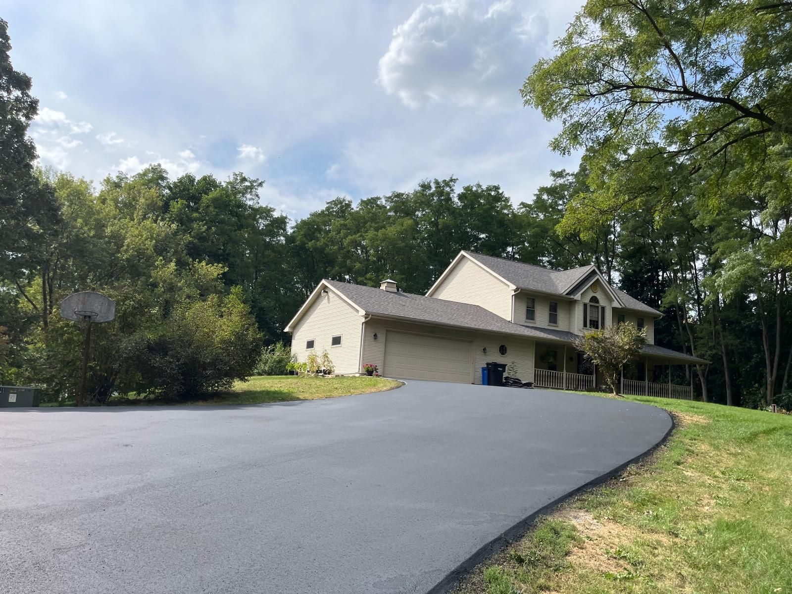 Asphalt driveway leading to a two-story beige house with a detached garage, set amongst trees, under a cloudy sky.