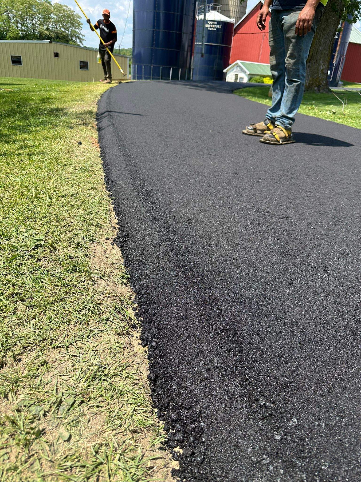 New asphalt path being laid; person working on path, another raking; green grass, silo and building background.
