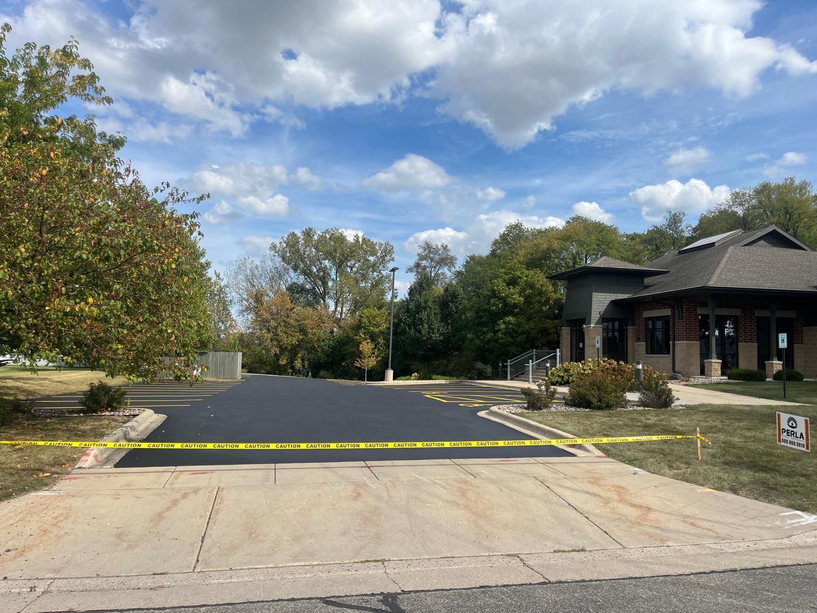 Freshly paved asphalt driveway with yellow caution tape, leading to a building under a cloudy sky.