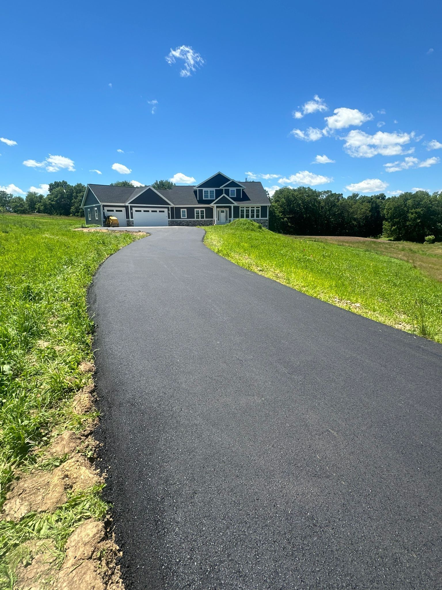 Paved driveway leads to a dark-colored house on a sunny day. Green fields surround the driveway.