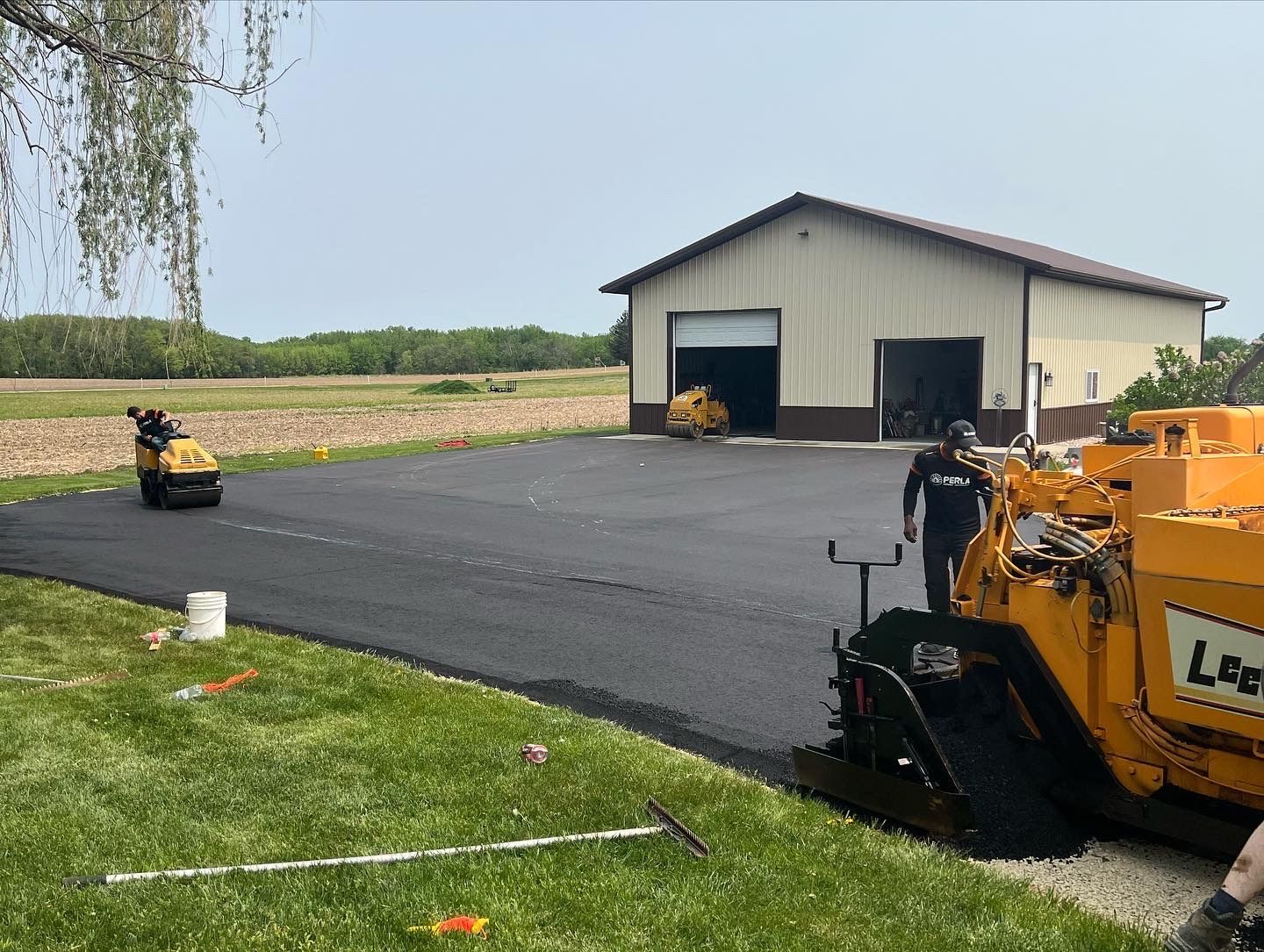 Asphalt paving in progress on driveway near a beige garage, with machinery and workers.