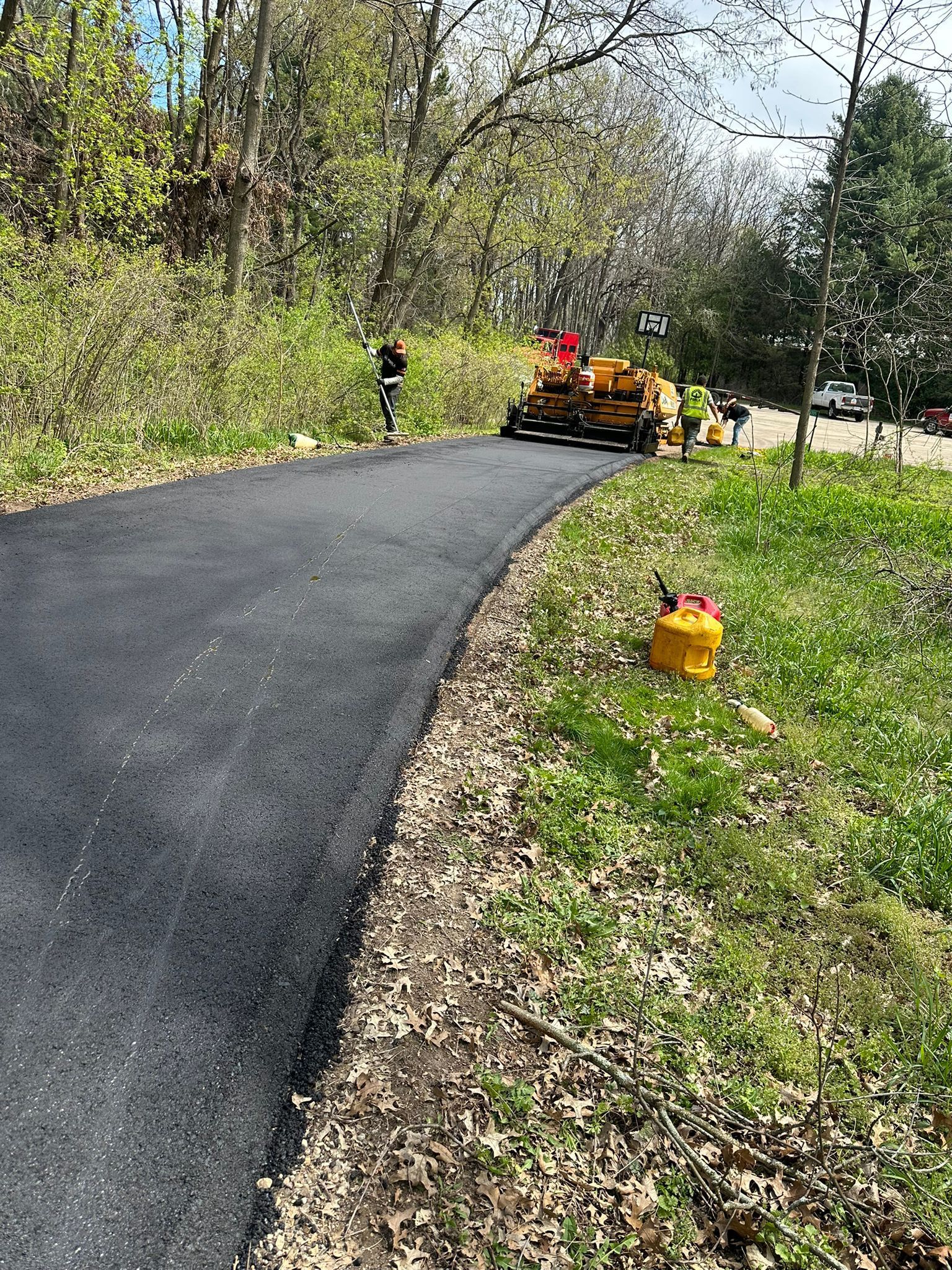 Paved asphalt path with construction equipment and workers, next to grassy area and trees.