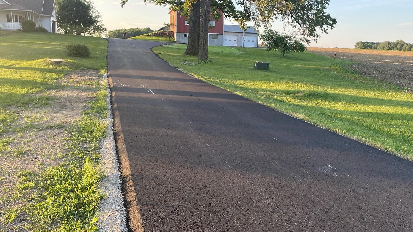 Paved driveway leading uphill towards a house and detached garage on a grassy rural property.