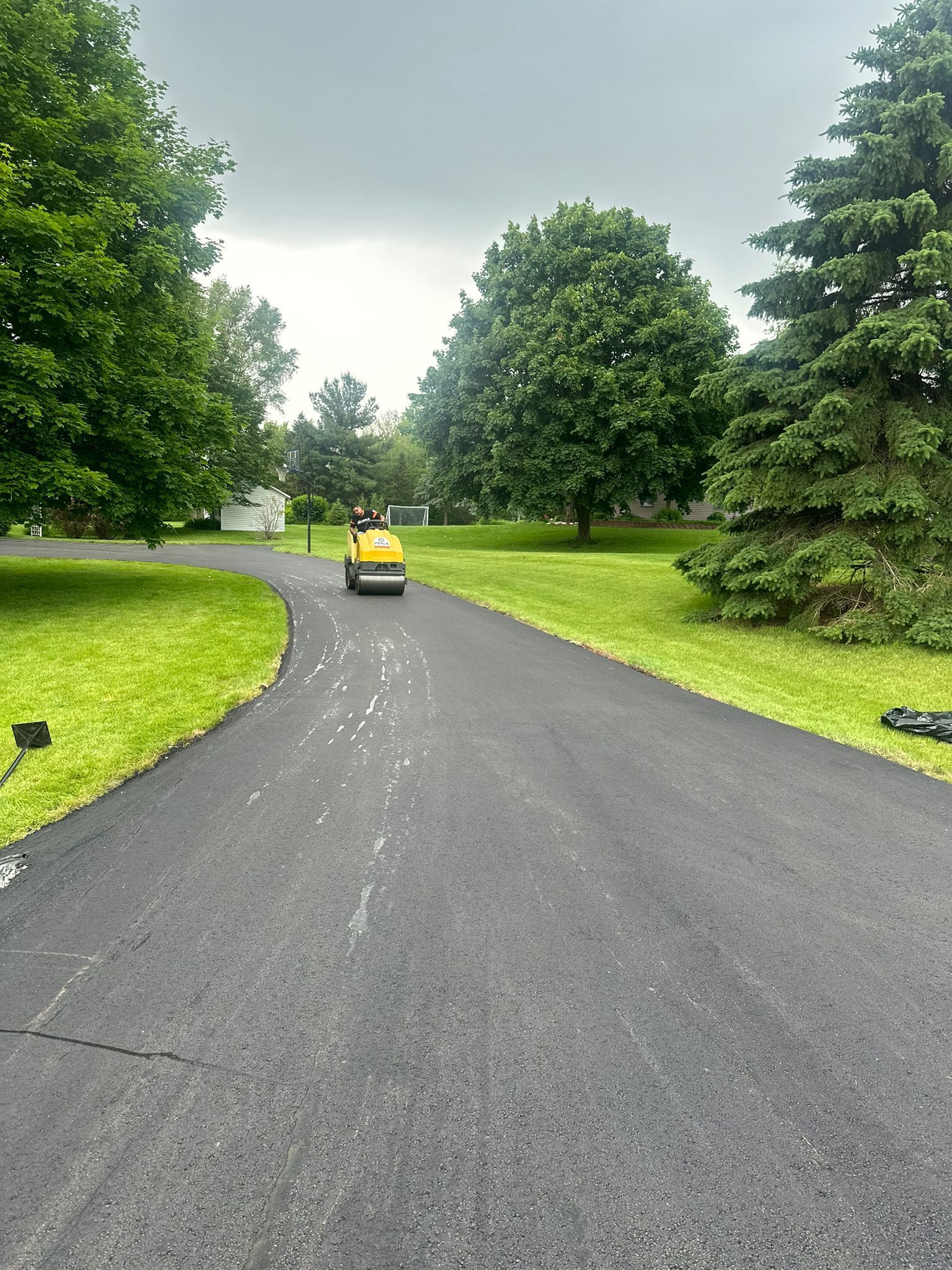 Asphalt driveway being rolled by a yellow machine, surrounded by green grass and trees under a cloudy sky.
