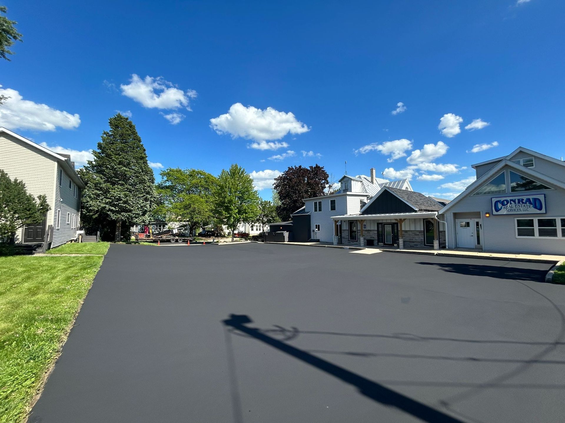 Black asphalt parking lot, blue sky, buildings, trees, and cloudy. The building on the right has 
