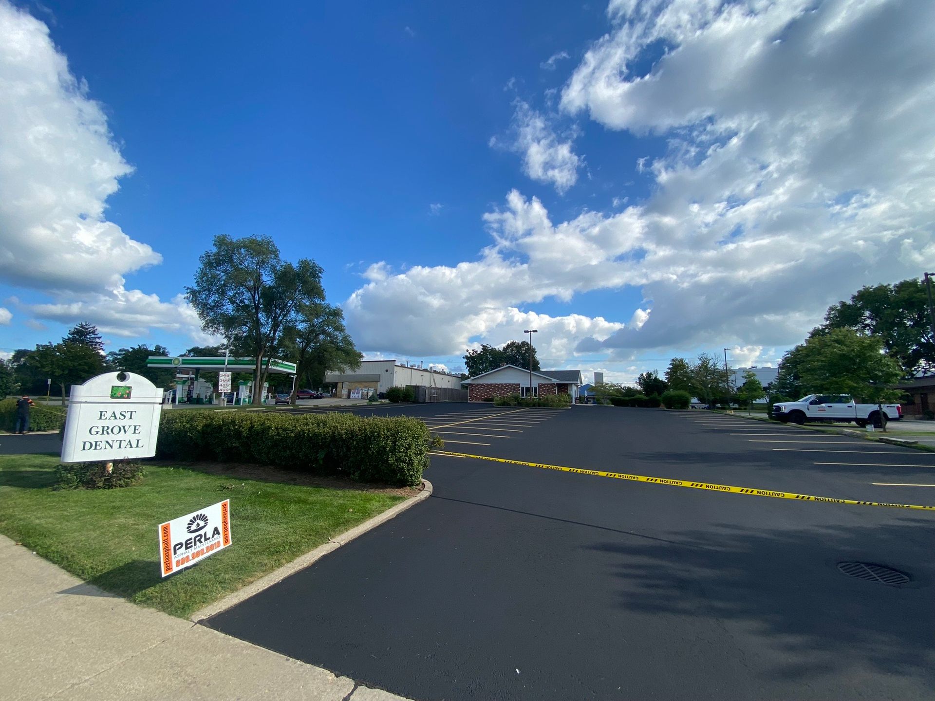 Sunny day over a paved lot with yellow caution tape, green bushes, and a sign. Buildings and blue sky in the background.