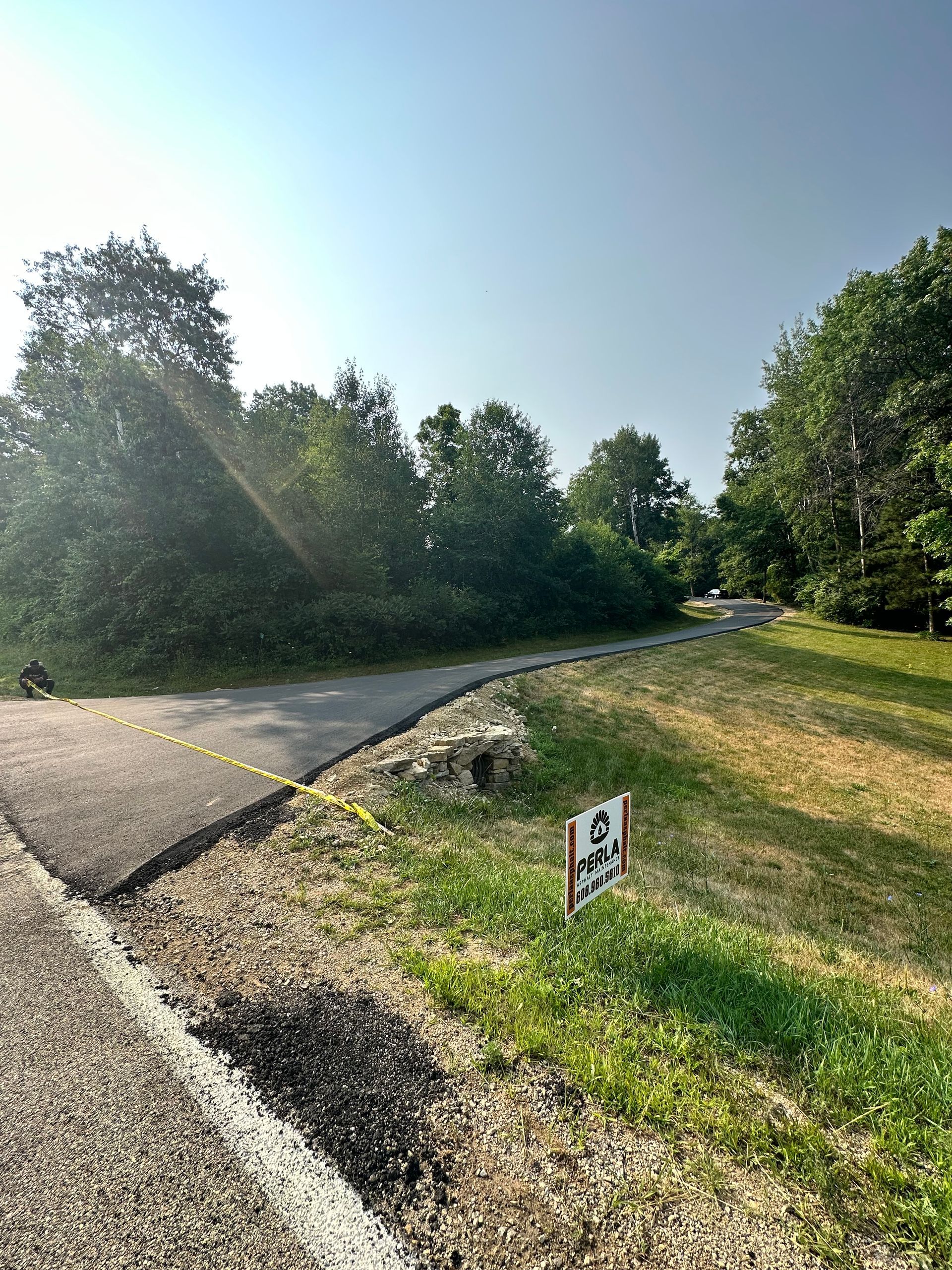Paved path curves into a tree-lined area, yellow caution tape, sign on grass. Bright sunny day.