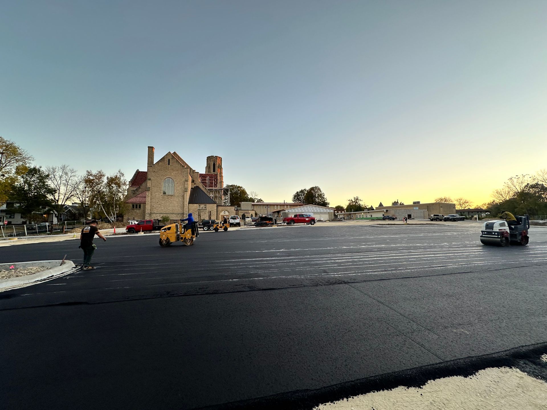 Asphalt paving on a large parking lot, with workers operating machinery and a church building in the background.