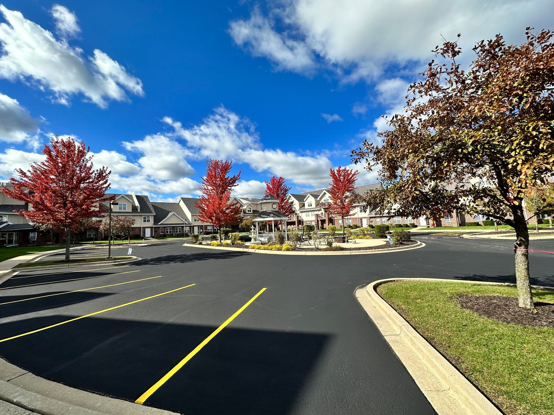 Parking lot with red-leafed trees, buildings, and a gazebo under a blue sky with clouds.